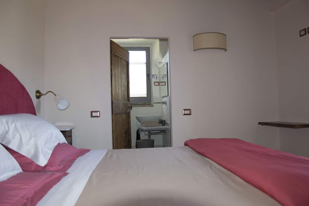 A bedroom with a bed, a bedside table, a mirror, a lamp, and a view into the bathroom.