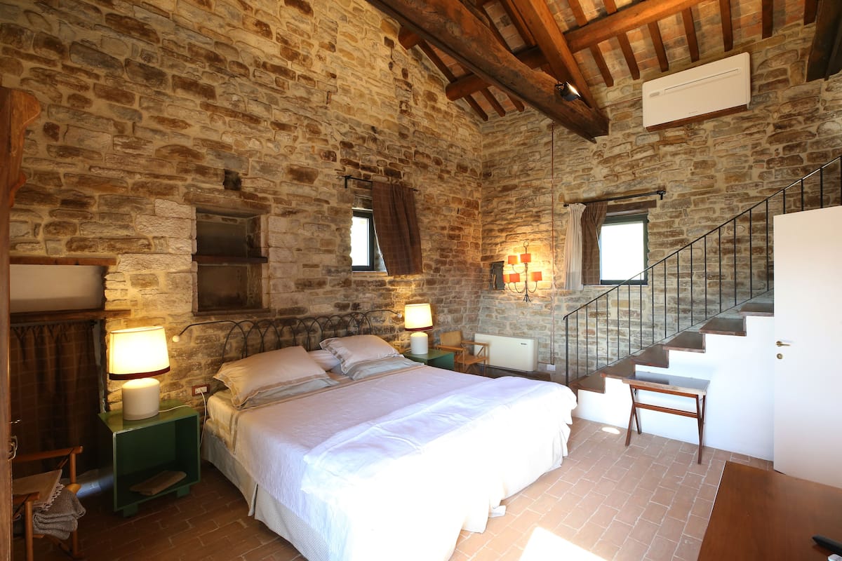 A bedroom with a stone wall, a wooden ceiling, a bed with white bedding, a nightstand with a lamp, a window with curtains, and a staircase.
