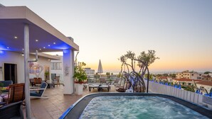 Een terras op het dak met een jacuzzi, lounge stoelen en uitzicht op de stad bij zonsondergang.