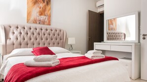 Een net opgemaakt bed met een rode deken, witte kussens en opgevouwen handdoeken. Daarboven hangt een grote spiegel, naast het bed een witte nachtkastje met een lamp, en een deur aan de rechterkant.