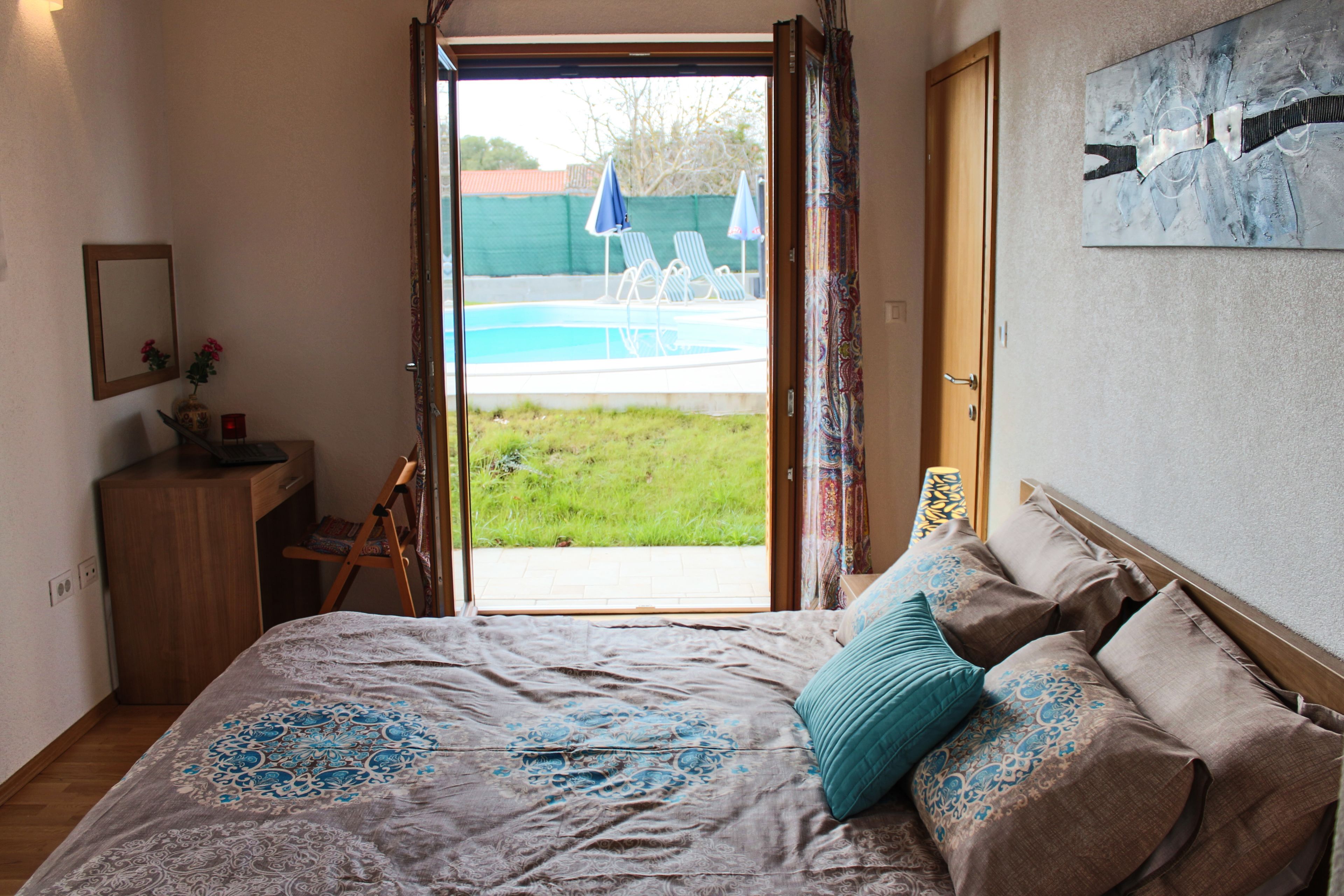A bedroom with a bed, a desk, a chair, a window with a view of a swimming pool, and a door.