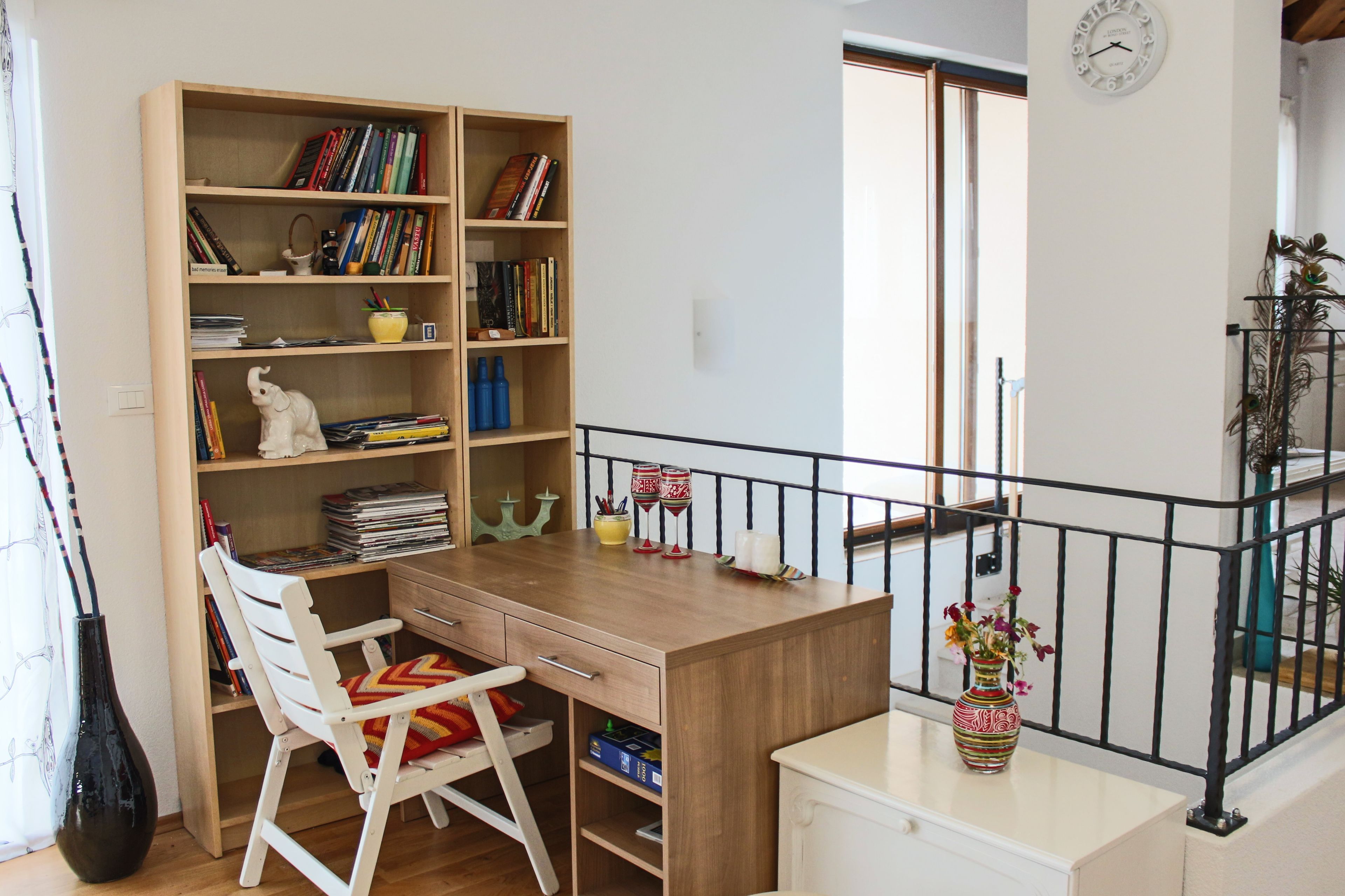 A study area with a wooden desk, white chair, bookshelf, and a small table with a vase.