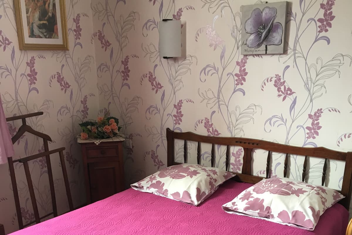 Une chambre à coucher avec un lit en bois, une literie rose, un papier peint à motifs floraux, une petite table de chevet ornée d’un vase de fleurs et un tableau encadré accroché au mur.