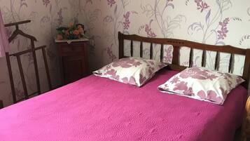 Un dormitorio con cama de madera, ropa de cama rosa, papel tapiz floral, una mesita de noche con un jarrón de flores y un cuadro en la pared.
