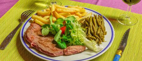 Een bord met eten, inclusief steak, frietjes, salade en bonen, met vork en mes op een groene tafelonderzetter.