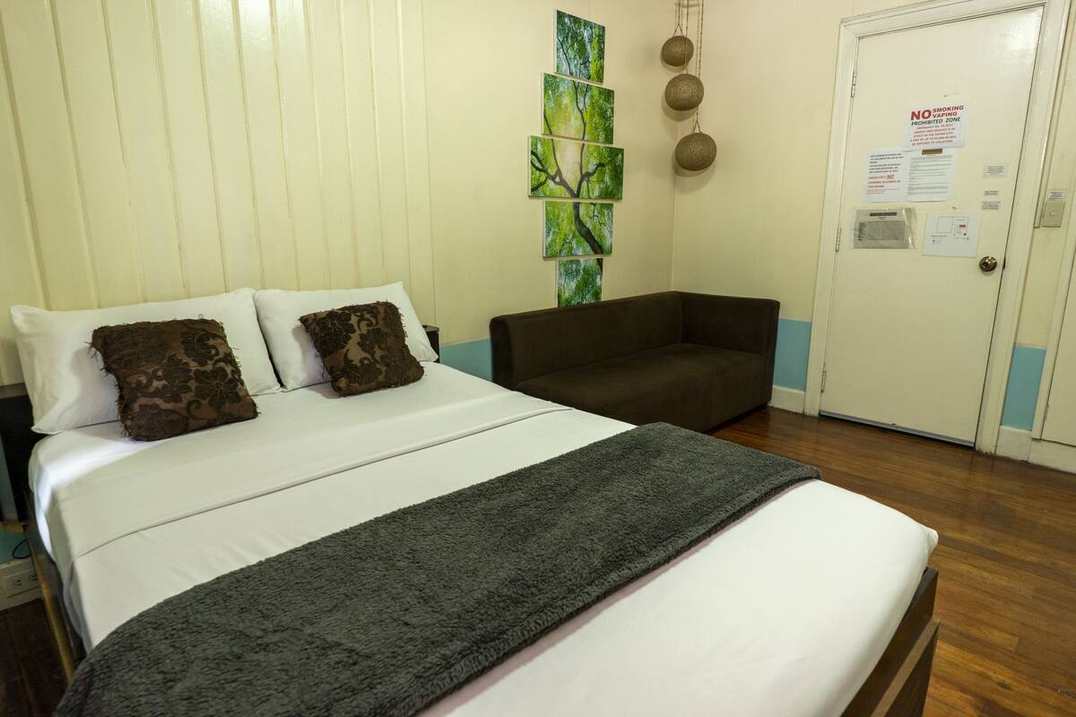 A hotel room with a bed, a brown sofa, a wall with a tree painting, and a door with a no-smoking sign.