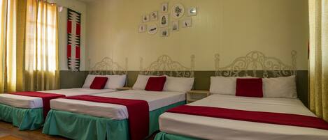 A hotel room with three beds, each with a red and white bedspread, and a green skirt. There are wall-mounted pictures and a patterned rug on the floor.
