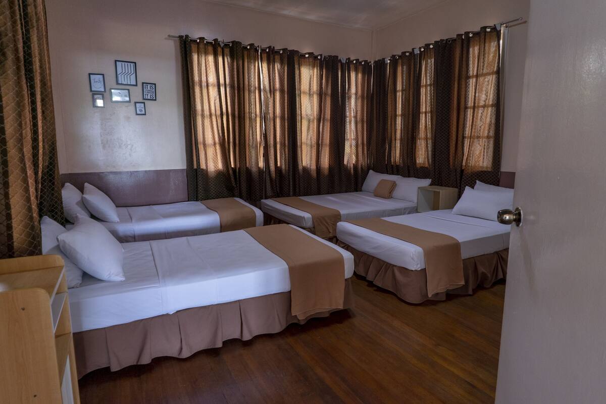 A hotel room with four single beds, a wooden floor, and curtains on the windows.