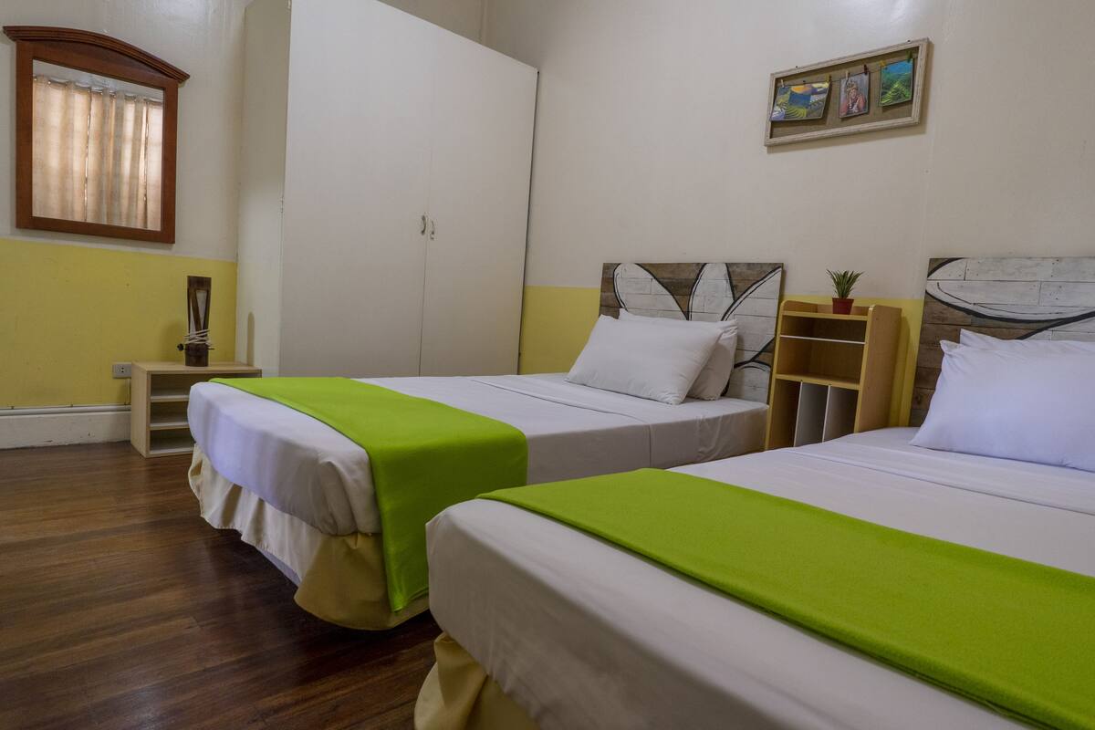 A hotel room with two beds, a wardrobe, a small table, and a shelf with a plant.