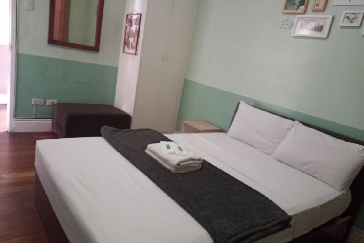 A bed with white linens, a dark grey blanket, and folded towels. There is a nightstand with a lamp, a small table, and a wall-mounted mirror. The room has a green wall and wooden flooring.