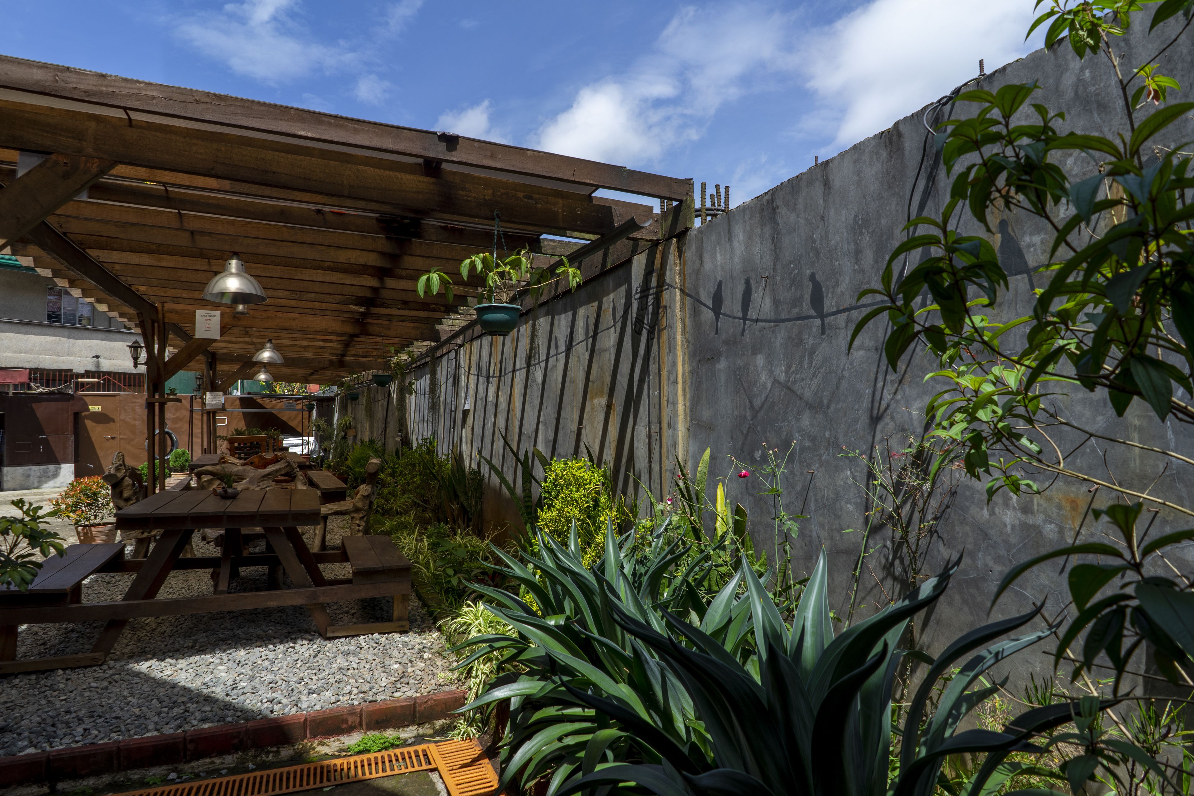 A covered patio area with a wooden table and benches, surrounded by potted plants and a concrete wall.