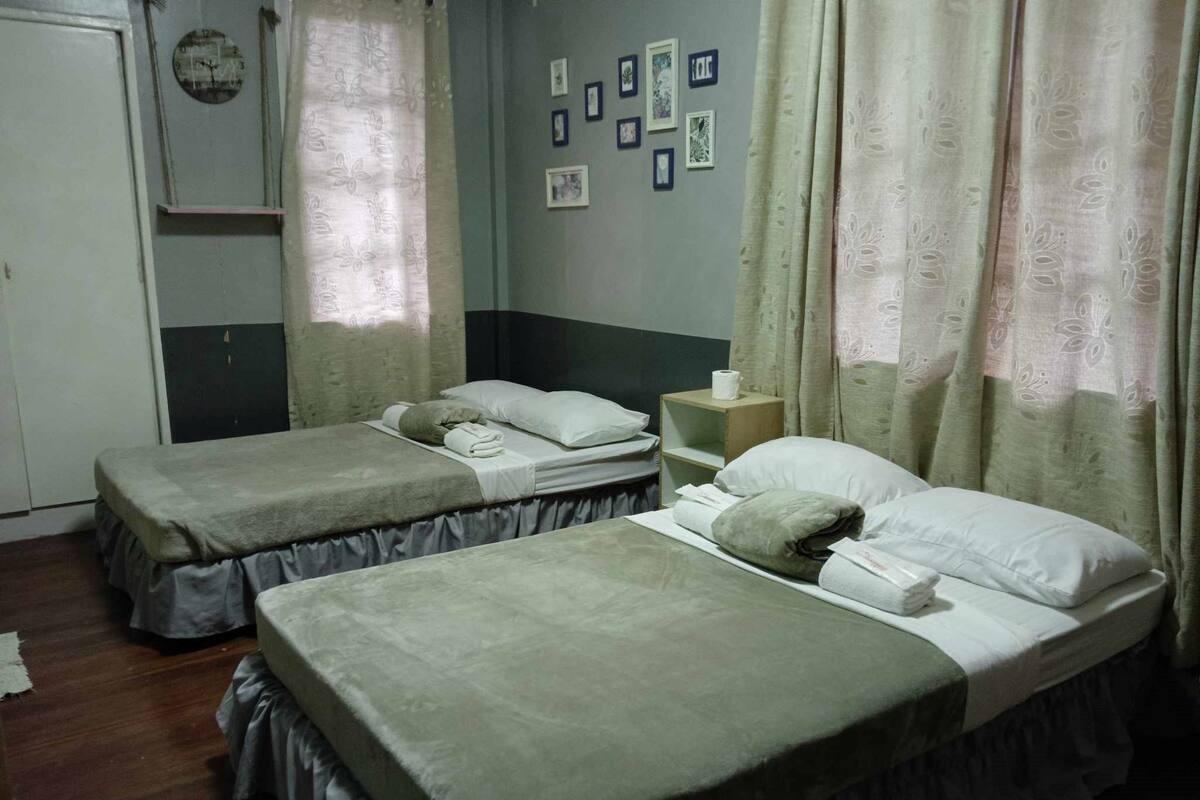 A room with two single beds, a wooden nightstand, a wall clock, and a shelf with framed pictures.