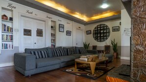 Una sala de estar con un sofá gris, una mesa de centro y una estantería. Hay cuadros enmarcados en la pared y un aviso en la puerta.