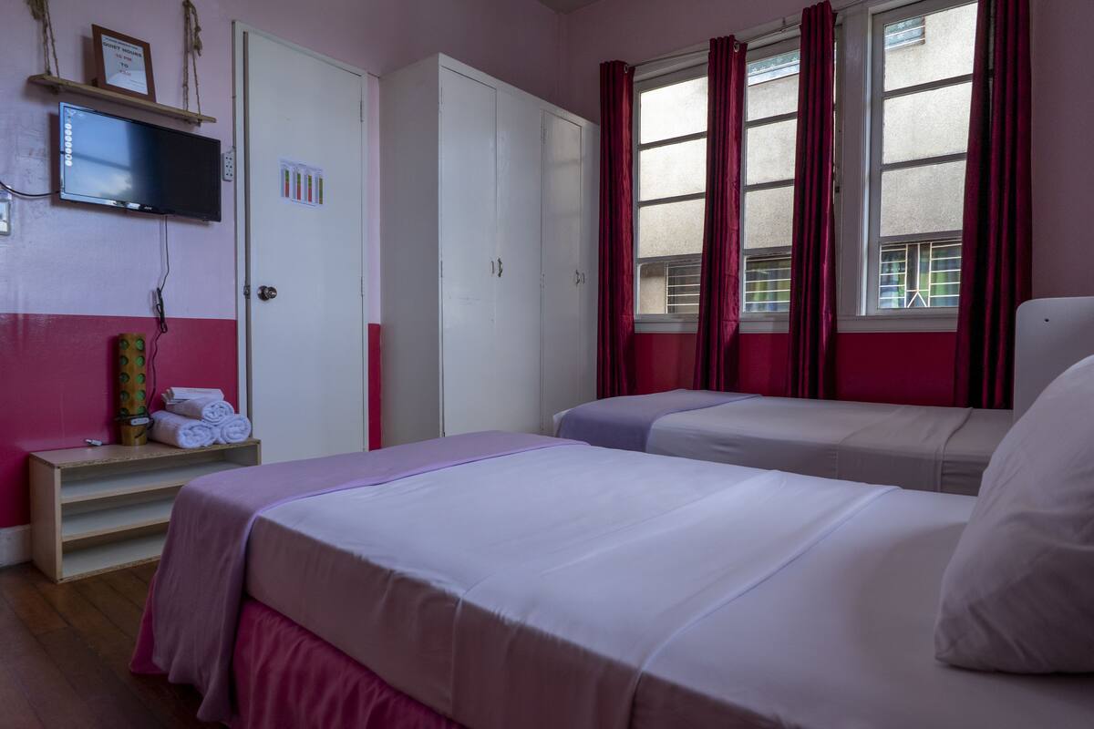 A hotel room with two beds, a TV mounted on the wall, a wardrobe, and a window with curtains.