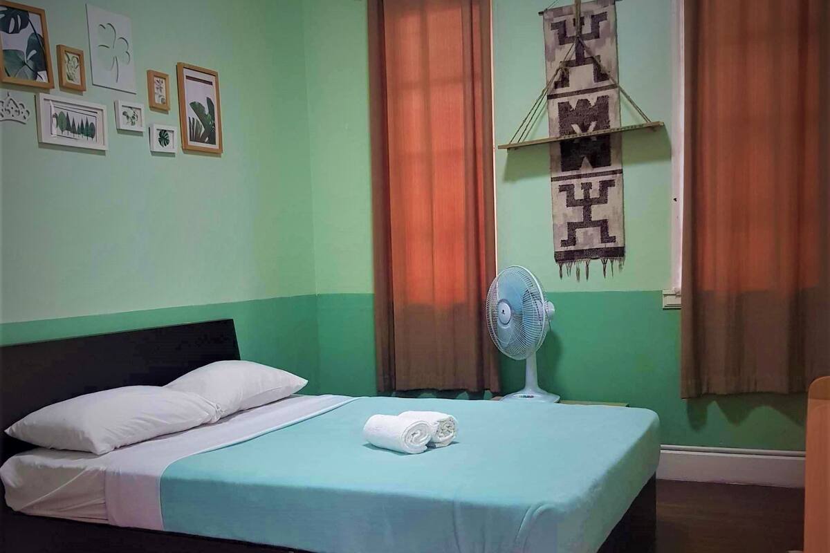 A neatly made bed with white pillows and a blue bedspread, a white fan on the floor, and a wall with framed pictures and a hanging sign.