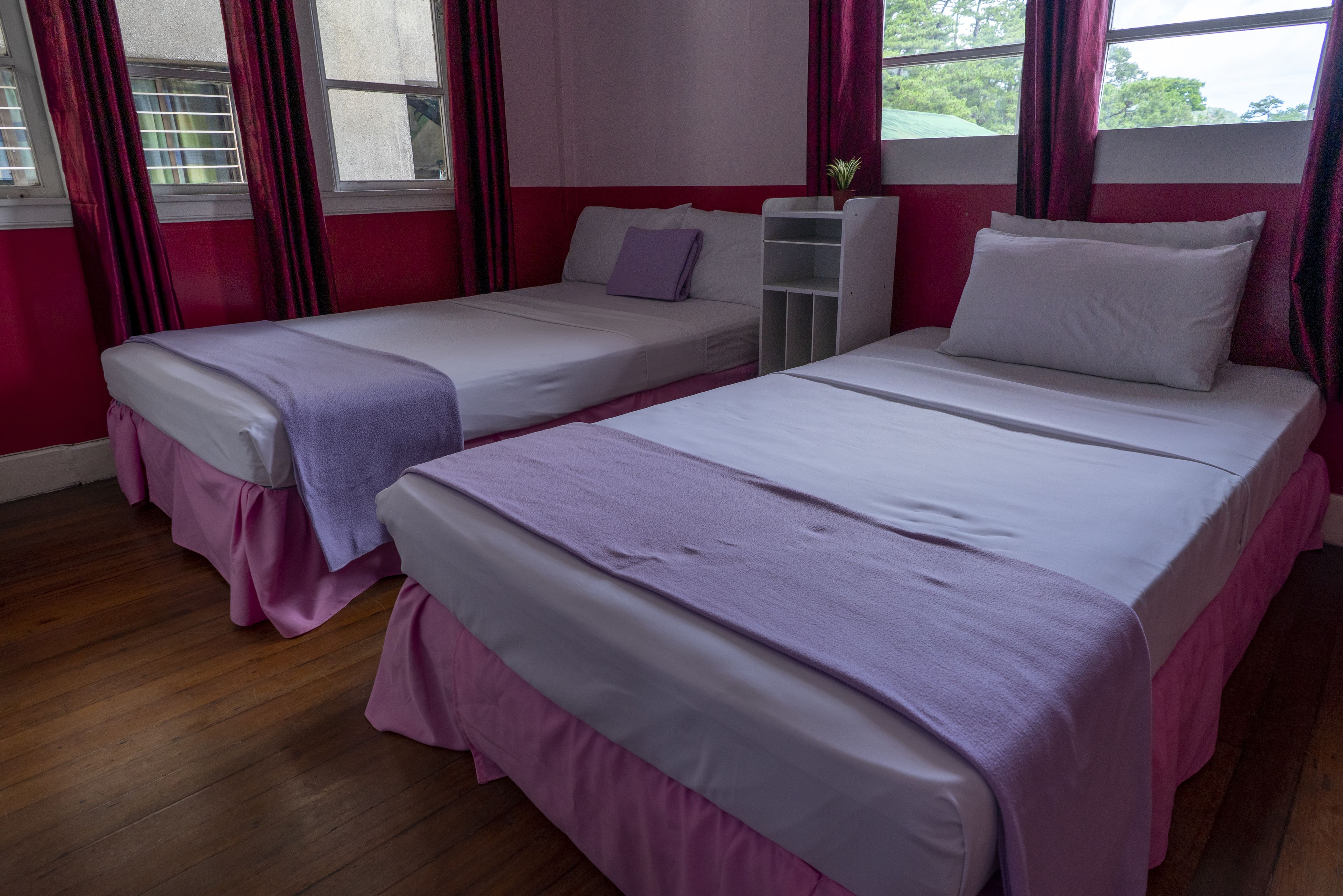 Une chambre d’hôtel avec deux lits simples, chacun agrémenté d’une jupe de lit rose et d’une couverture violette. Une table de chevet blanche se trouve entre les lits, et une fenêtre est ornée de rideaux rouges.