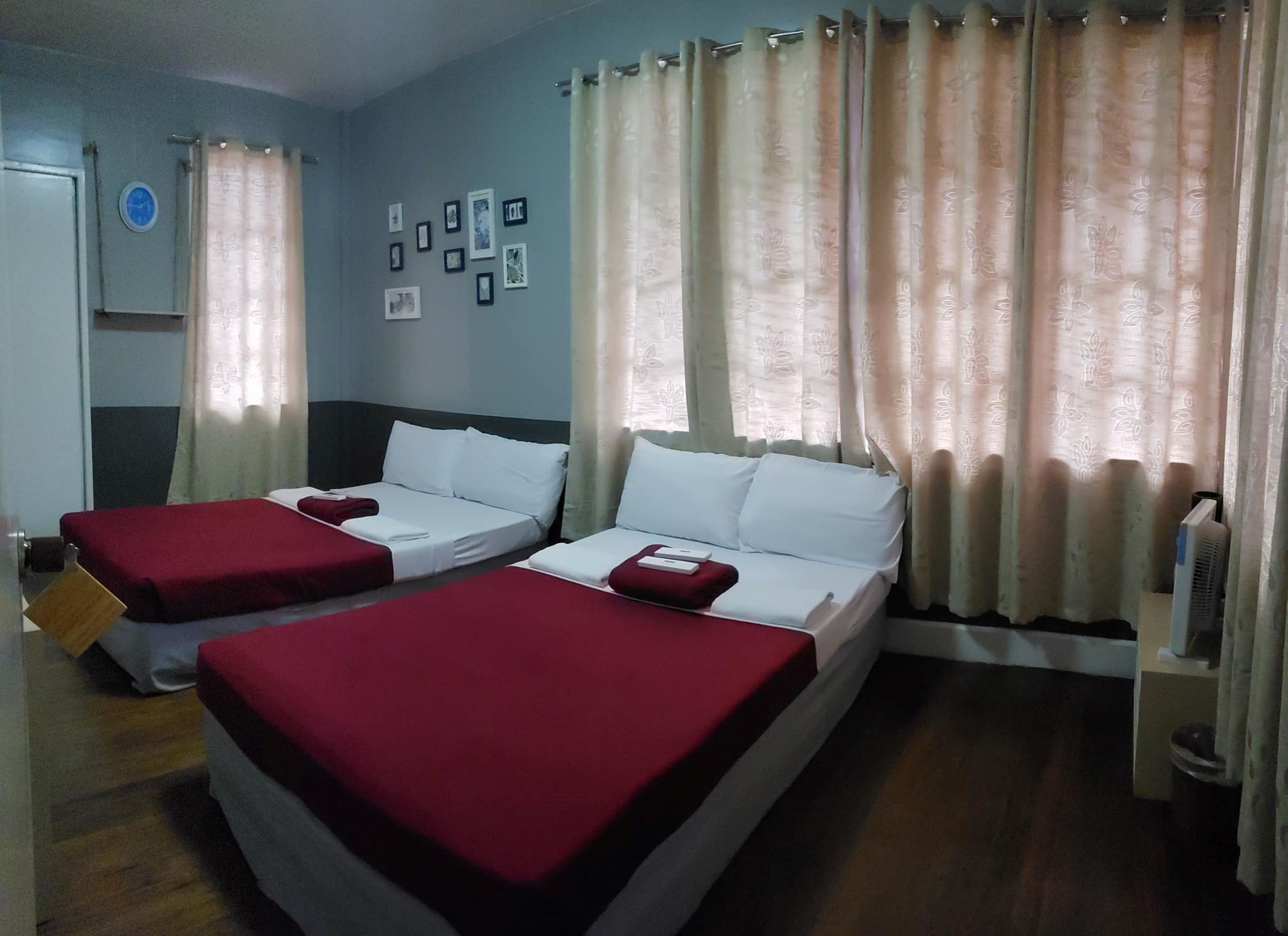 Una camera d'albergo con due letti, un tavolino, una sedia, una sveglia appesa al muro e una finestra con tende.