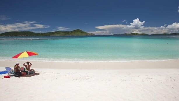 Une plage de sable blanc bordée d’une eau turquoise et cristalline. Un parasol rouge et jaune offre de l’ombre.