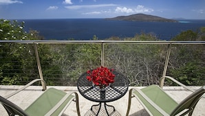 Un balcon avec une table et des chaises, offrant une vue panoramique sur l’océan et une île lointaine.