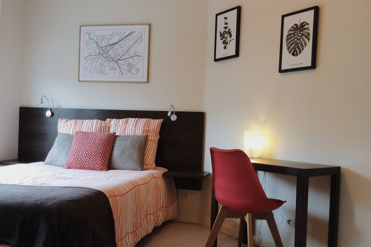 A bedroom with a bed, a desk, and two framed botanical prints on the wall.