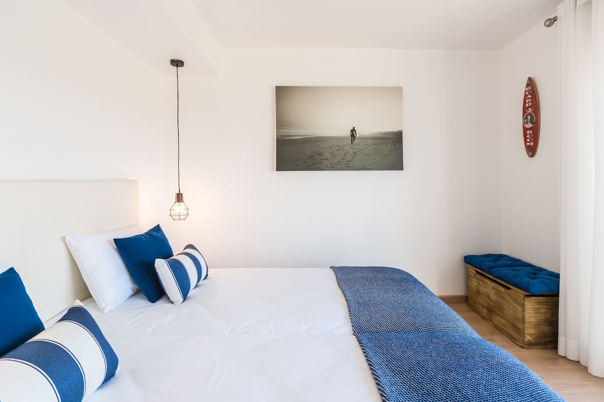 A bedroom with a large bed, a wooden bench, a wall-mounted picture, and a hanging light fixture.