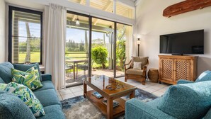A spacious living room with a large sliding glass door leading to a patio, a comfortable sofa set, a wooden coffee table, and a television on a wooden cabinet. - Spacious Townhome with Golf Course Views (Kahuku)