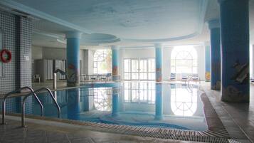 An indoor swimming pool with clear water, surrounded by blue-tiled walls and columns, and large windows allowing natural light.