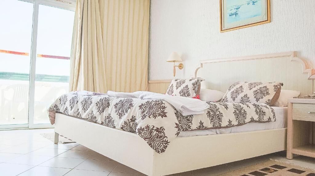 A hotel room with a bed, a nightstand, a lamp, a window with curtains, and a painting on the wall.
