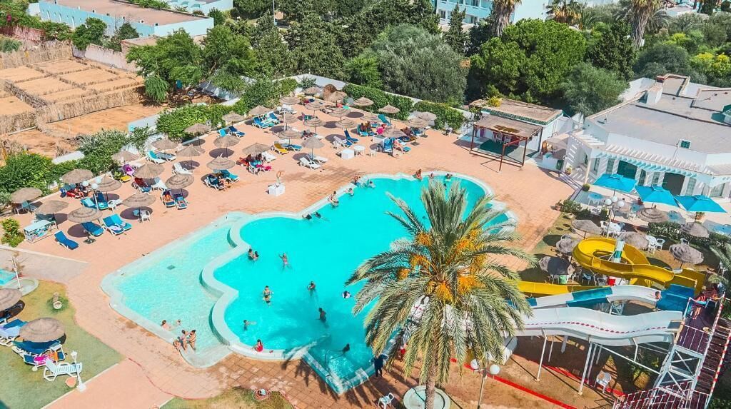 An aerial view of a resort with a swimming pool, sun loungers, umbrellas, and a water slide.
