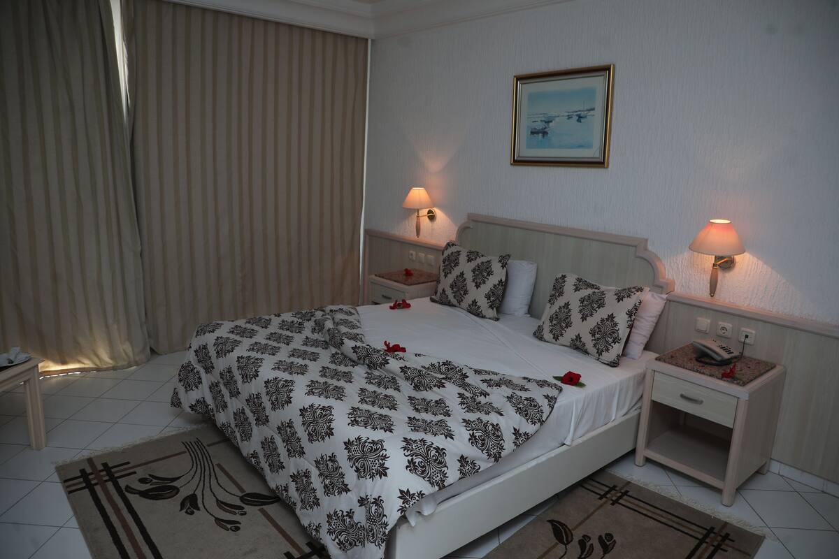 A neatly made bed with patterned bedding, two bedside tables with lamps, a framed picture on the wall, and a window with curtains.