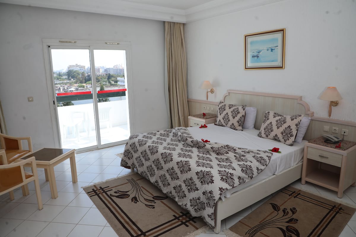 A hotel room with a bed, two bedside tables, a chair, a window with a view of buildings, and a door.