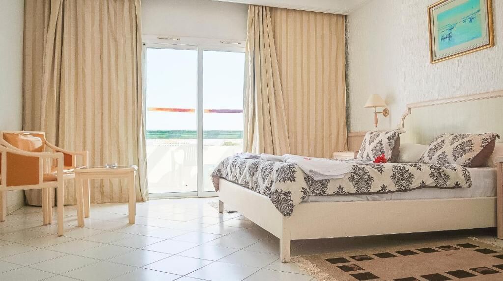 Habitación de hotel con cama, una silla, una mesa y vista a la playa a través de una puerta corrediza.