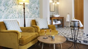 A living room with a mustard sofa, two pillows, a floor lamp, a round coffee table with a bowl and glasses, and a bathroom with a sink, mirror, and towel rack. - The Old New Inn (Cheltenham)