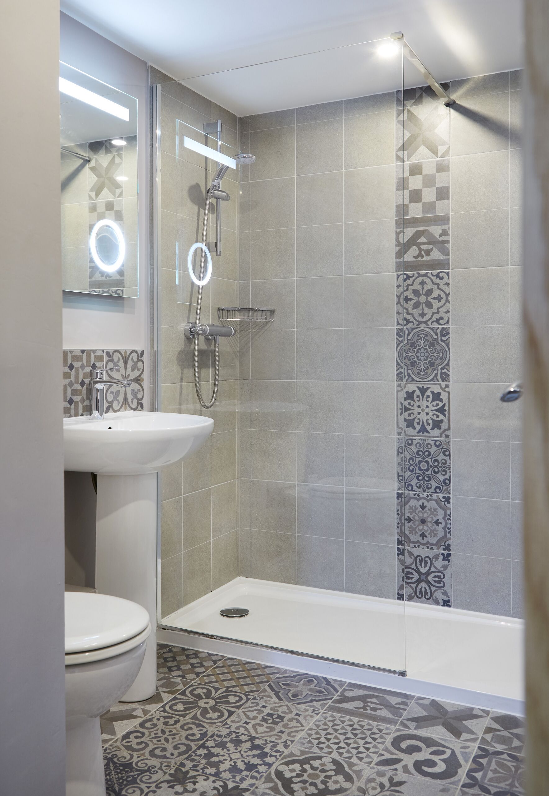 Et badeværelse med toilet, håndvask, brusebad og et gulv med mønstret flisearbejde.
