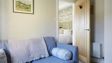 A room with a blue sofa, a herringbone-patterned throw blanket, and a plaid pillow. A wooden door with a black handle is visible, leading to a bedroom with a bed and a radiator.