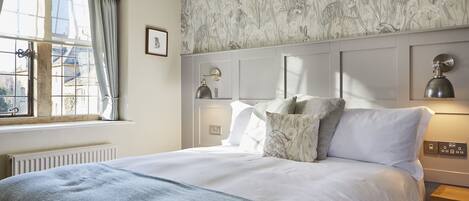A neatly made bed with a grey blanket, a window with curtains, a wall with a floral pattern, and a bedside table with a lamp.