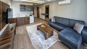 Un salon moderne avec un canapé gris, une table basse en bois et un coin cuisine doté de meubles en bois et d’un réfrigérateur encastré.