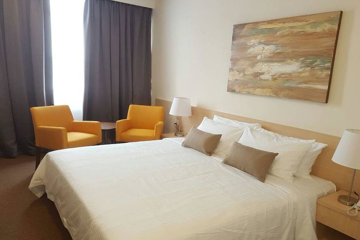 A hotel room with a large bed, two chairs, a painting on the wall, and a nightstand with a lamp.