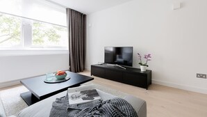 A modern living room with a flat-screen TV on a black TV stand, a dining table with a bowl of fruit, a magazine, and a potted orchid. - Craven Hill by Lime Street (London)