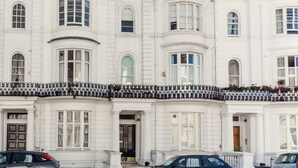 Une rangée de bâtiments blancs, dotés de nombreuses fenêtres et de balcons, une rue devant et des voitures garées.
