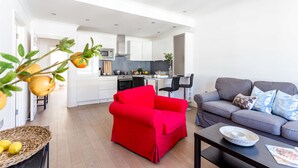 Un salon moderne avec un fauteuil rouge, un canapé gris et une table basse. On aperçoit une cuisine à l’arrière-plan, avec des meubles blancs et un four à micro-ondes.