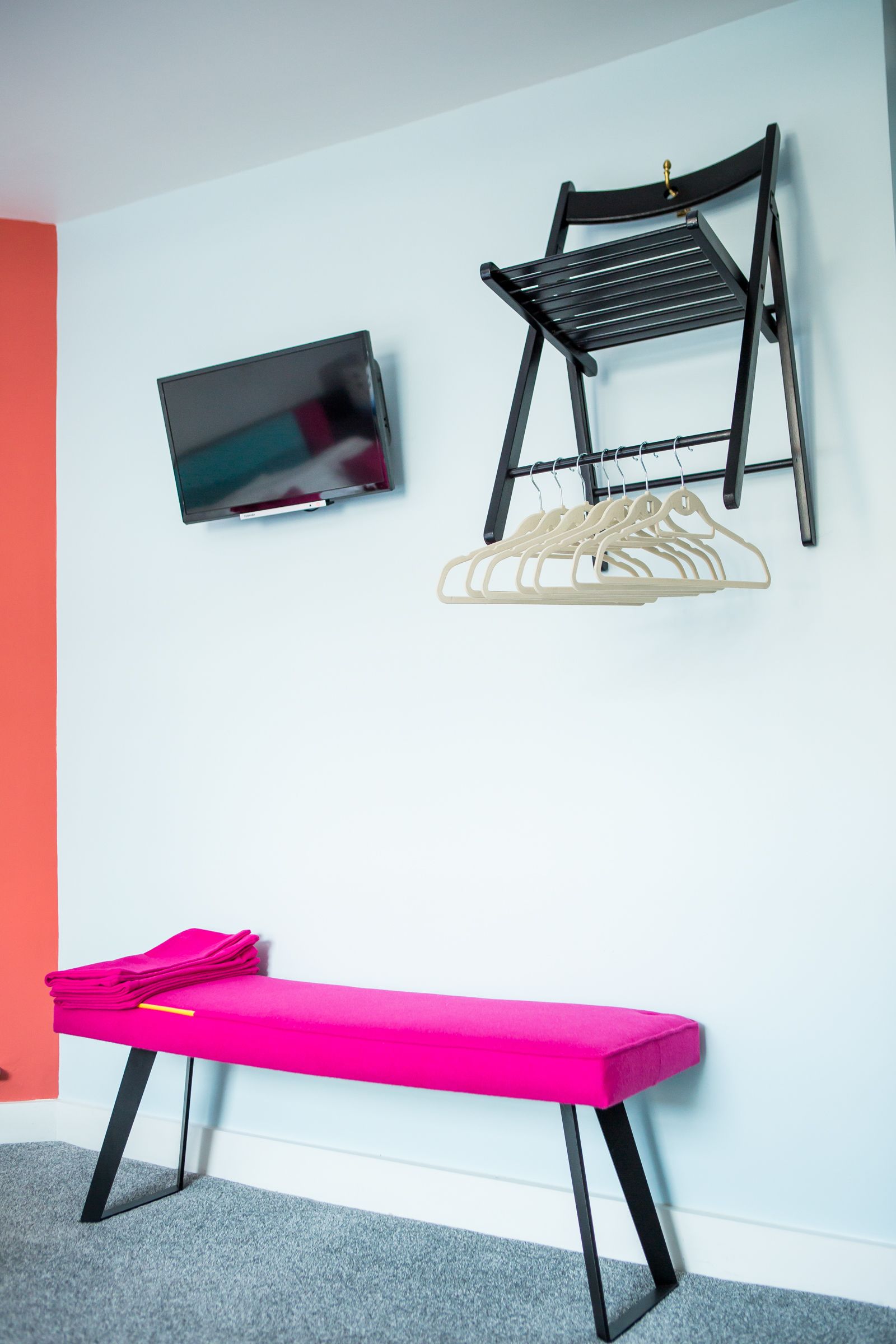 A room with a TV mounted on the wall, a black chair with a shelf, a bench with pink cushions, and a grey carpet.