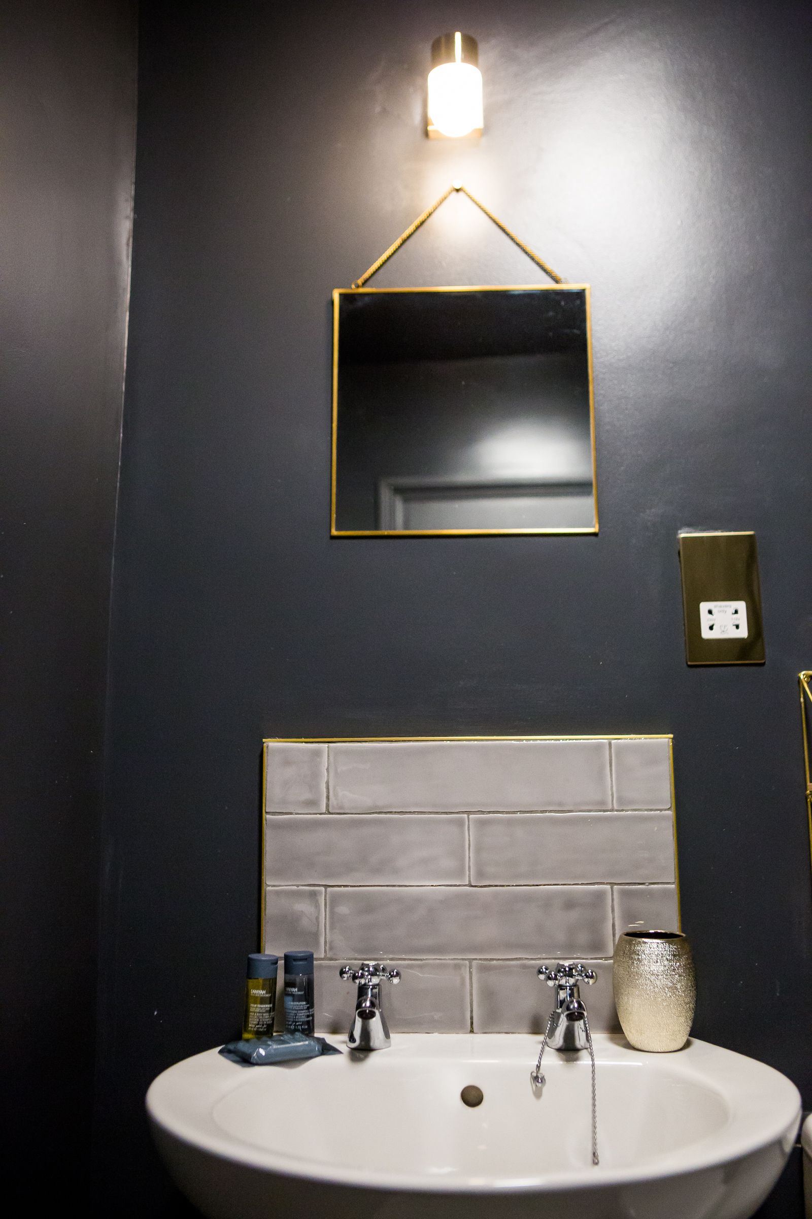A bathroom with a white sink, two faucets, a mirror with a gold frame, a light fixture above the mirror, and a tiled wall.