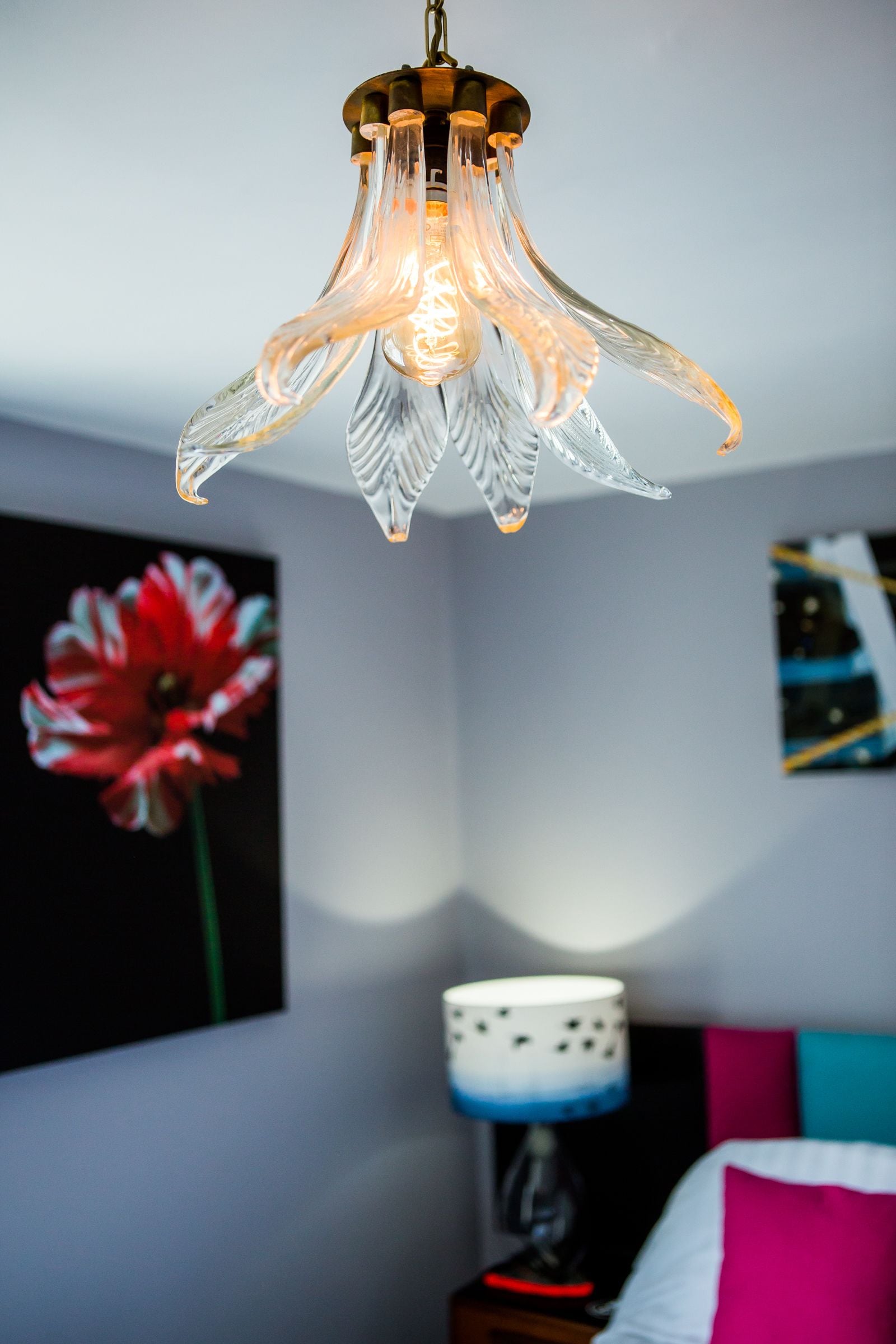 A room with a hanging glass light fixture, a red flower painting, a bedside lamp, and a bed with a pink pillow.