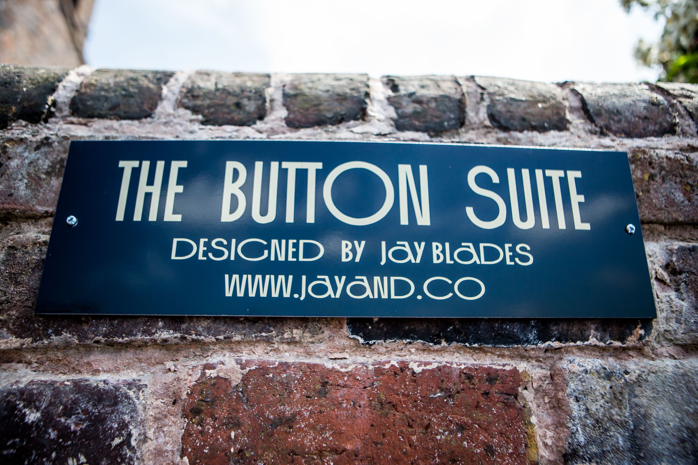 Tiiliseinään on kiinnitetty kyltti, jossa lukee: ”The Button Suite – Jay Bladesin suunnittelema.”