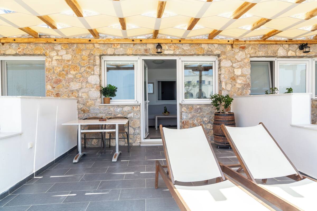 Un espacio al aire libre techado con paredes de piedra, una mesa, sillas y plantas en macetas.