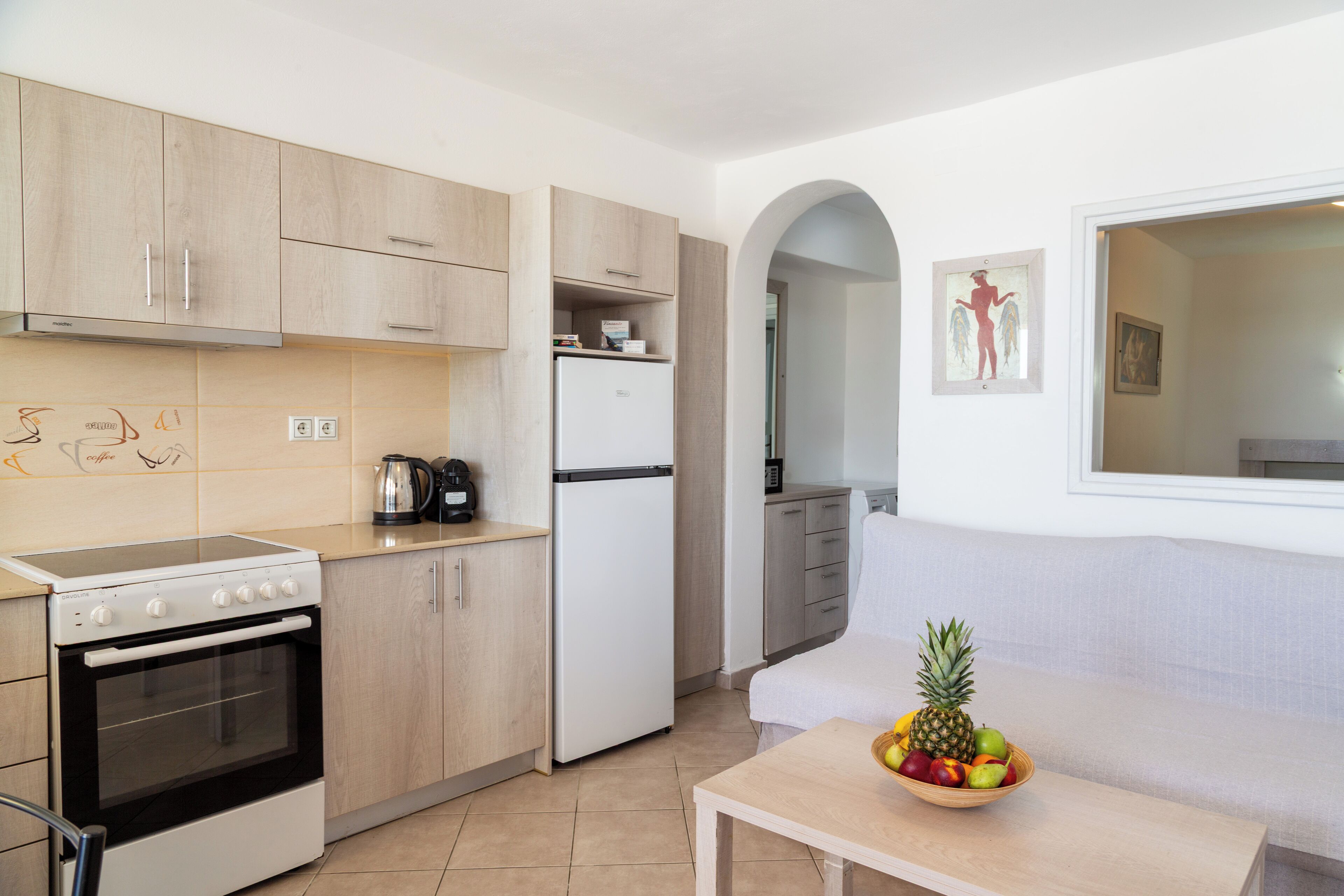 A modern kitchen with wooden cabinets, a white stove, and a refrigerator. There is a fruit bowl on a table and a couch in the adjacent living area.