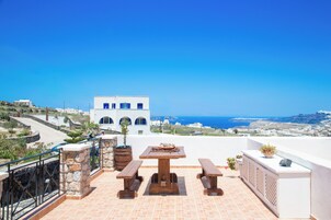 Una terraza con mesa y bancos de madera, con vista a un paisaje costero con edificios blancos y el mar.