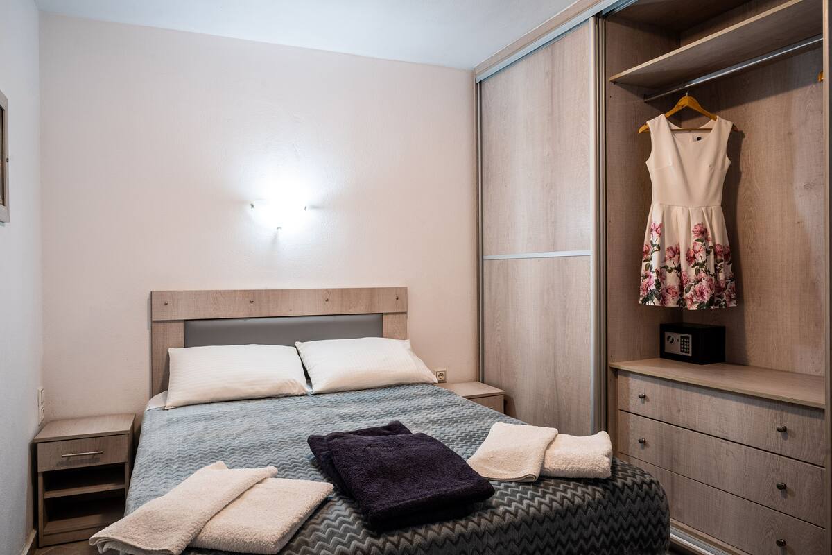 Una cama bien hecha con almohadas y una colcha, una mesita de noche con lámpara, un armario y un vestido colgado.