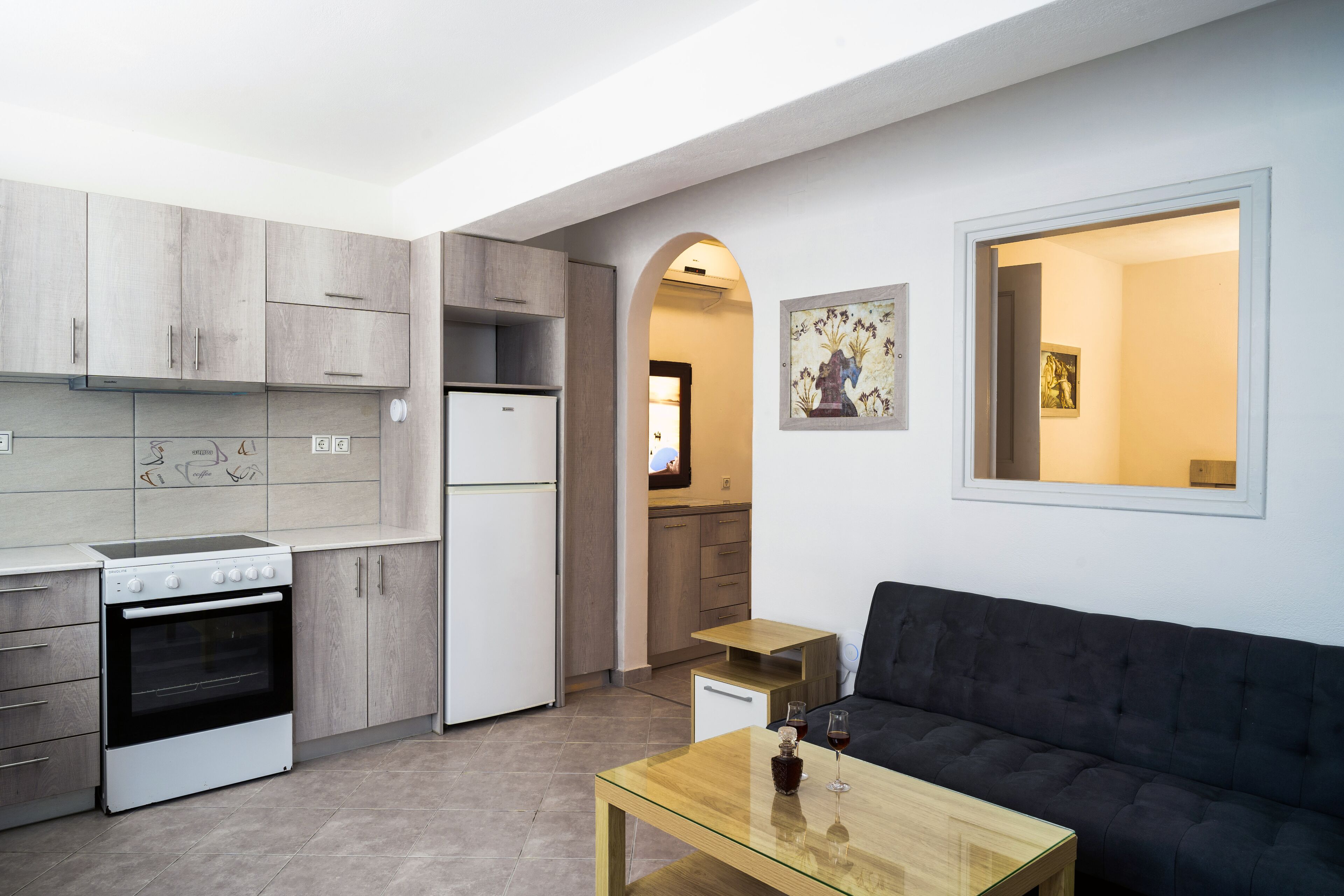 A compact kitchen with a stove, refrigerator, and wooden cabinets. There is a dark sofa and a small table with a glass top.